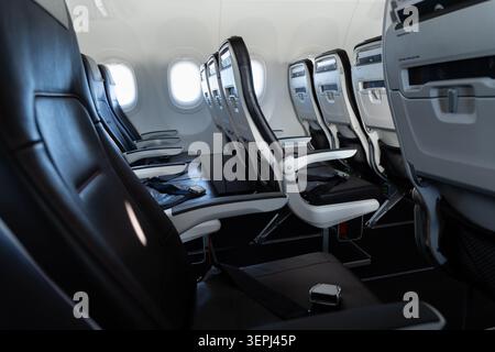 Reihen unbesetzter Flugzeugsitze mit Sicherheitsgurten, Kopfstützen und Tablettentischen, die in einer modernen, schmalkörnigen Flugzeugkabine sichtbar sind Stockfoto