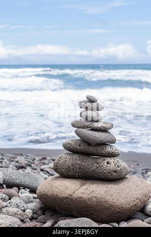 Ausgeglichener vulkanischer Steinkairn an einem steinigen Strand mit Wellen des Pazifischen Ozeans im Hintergrund auf Hawaiis Big Island, symbolisiert Gleichgewicht, Achtsamkeit und . Stockfoto