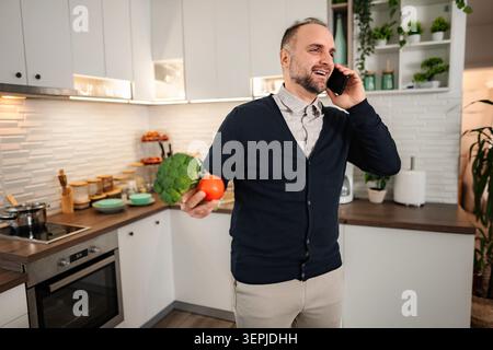 Mann, der am Telefon spricht und Gemüse in der Küche hält, lächelnd Stockfoto
