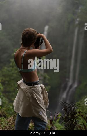 Frau in Crop Top fotografiert Wasserfall im nebeligen Dschungel, Bali Stockfoto