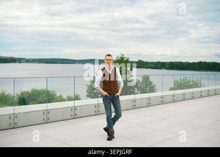 Mann mittleren Alters steht auf einem modernen Balkon mit Blick auf See und Bäume. Wasser reflektiert Wolken darüber. Der Mann scheint entspannt zu sein, während er legere Kleidung trägt Stockfoto