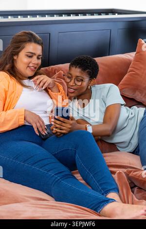 Verschiedene Freundinnen sitzen dicht auf rostfarbenen Sofas und halten ein dunkelblaues Smartphone. Frauen, Paar, Zuhause, Terrakotta, gemütlich, Wärme, Mitgefühl Stockfoto
