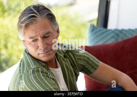 Senior Mann sitzt auf der Couch, in gestreiftem Hemd, blickt durch das Fenster, rote und türkisfarbene Kissen. Besinnlich, ruhig, sonnendurchflutete Zimmer, Wohnzimmer, Sofa, gemustert, grau Stockfoto
