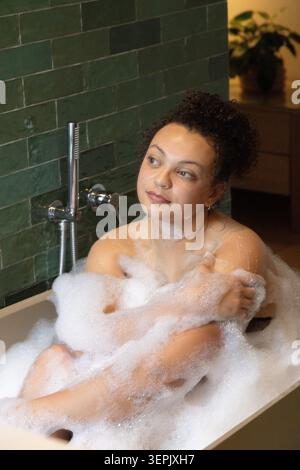 Afroamerikanische Frau sitzt in freistehender Wanne vor dunkelgrünen Fliesen, bedeckt mit weißem Schaum. Badewanne, Wellness, minimalistisch, Badezimmer, Innenausstattung, ruhig, besinnlich Stockfoto