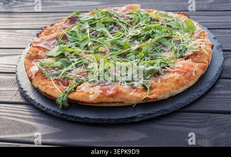 Frisch gebackene Pizza mit Schinken, Rucola und Käse, serviert auf einem rustikalen Holztisch. Stockfoto
