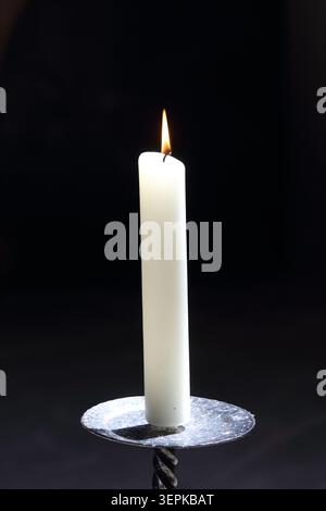 Vertikale Aufnahme einer einzelnen beleuchteten weißen Kerze mit einer hellen goldenen Flamme in einem Metallhalter vor schwarzem Hintergrund Stockfoto