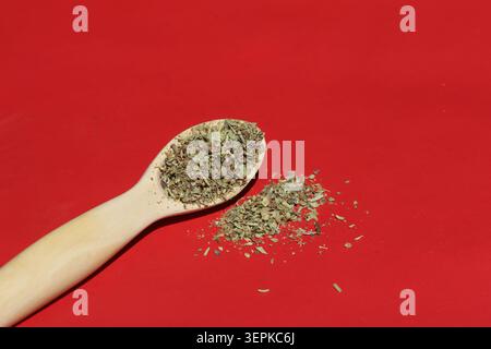 Abgewinkelte Nahaufnahme getrockneter, zerstoßener Oregano-Kräuter auf einem Holzlöffel mit verstreuten Flocken auf einem soliden roten Hintergrund Stockfoto