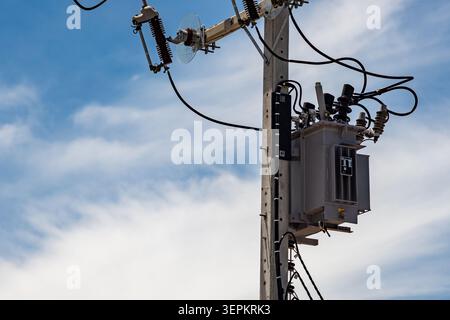 Nahaufnahme eines Versorgungsmasts im Freien mit Stromtransformator und Drähten, die die Infrastruktur der Energieverteilung vor einem hellblauen s hervorheben Stockfoto
