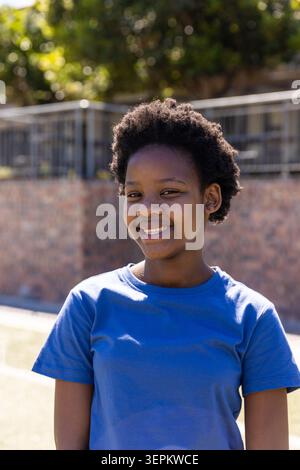 Teenager Afroamerikanerin lächelt, steht im sonnigen Park mit blauem T-Stück an Ziegelgelgeländer Stockfoto
