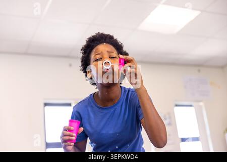 Afroamerikanischer Schuljunge, der Seifenblase mit rosa Becher und weißem Zauberstab im Klassenzimmer bläst Stockfoto
