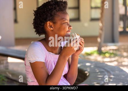 Afroamerikanisches Kind sitzt in rosa Kleidern auf der Schulbank, hält Sandwich und lächelt Stockfoto