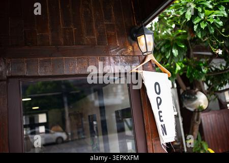 Eine gemütliche Szene im Freien mit einem Café-Eingang mit einer Holzfassade, üppigem Grün, einem einladenden „Open“-Schild und einem einladenden Ambiente, das perfekt für zwangloses Ambiente ist Stockfoto