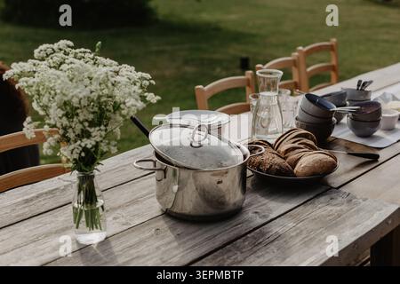 Einladendes Essen im Freien auf einem rustikalen Holztisch mit frischem Brot, Kochgeschirr, Gläsern und Wildblumen. Perfekt für Themen der Gastfreundschaft, Stockfoto