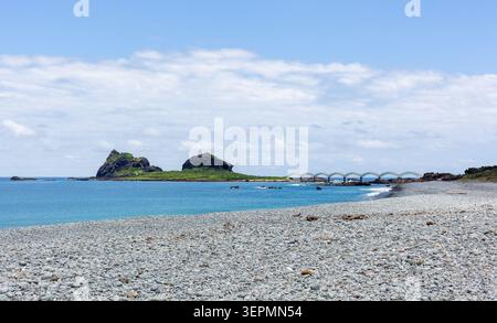 Die Sanxiantai Bridge verbindet die Insel mit dem Festland. Türkisfarbenes Wasser trifft auf Kiesstrand unter blauem Himmel. Grüne Vegetation bedeckt felsiges Gelände. Stockfoto