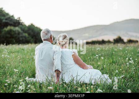 Glückliches Seniorenpaar, das auf Gras sitzt und den Sonnenuntergang ansieht. Stockfoto