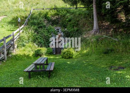 Ein in den Hang gebautes Gewölbe mit einem schmiedeeisernen Gittertor steht an der Rettenbachstraße. Davor lädt ein hölzerner Picknicktisch am Waldrand zu einer Pause ein. Rettenbachstraße, Spielbichl, Schattberg, Mittersill, Bezirk Zell am See, Salzburg, 5730, Österreich Stockfoto