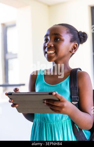 Afroamerikanisches Kind lächelt und hält Tablette in Folio, trägt in der Schule ärmelloses Kleid in blauem Blau Stockfoto