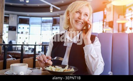 Kaukasische Ältere Geschäftsfrau Lächelt Am Telefon, Während Sie Salat Isst Und Kaffee Am Sonnigen Cafe Booth Trinkt, Stift Hält Und Notizen Schreibt, Entspannt Stockfoto