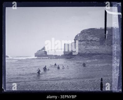 Pont Vieux überspannt im Oktober 1891 die Küstengewässer von Biarritz neben Rocher de la Vierge, wobei die Wellen des Atlantiks gegen die baskische Küste brechen, während ein Mann in der Nähe der Brücke mit Blick auf felsige Ausläufer und Surfen entlang der Küste des Resorts steht. Stockfoto