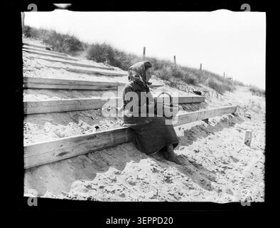 Eine junge Frau sitzt ruhig an einem Sandstrand in einer Küstenlandschaft und bietet einen ruhigen Moment an der Küste in Frankreich. Die Komposition spiegelt die Küstenerholung des 19. Jahrhunderts und das entspannte Leben im Freien wider, das aus einem Archivbild von 1880 erhalten wurde. Stockfoto