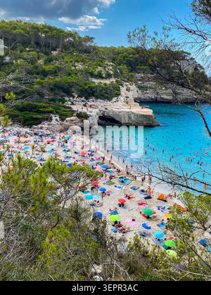 Blick aus einem hohen Winkel auf den Strand von Cala Mitjana auf Menorca, Balearen, Spanien. Wunderschöne unberührte Bucht mit feinem weißen Sand, voll mit Touristen, Kristall C Stockfoto