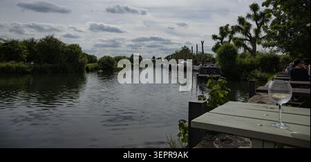 Ein Glas Gin Tonic auf einem Pub-Tisch mit Blick auf die Themse bei Lechlade mit Schmalbooten, die entlang des Ufers in der britischen Landschaft vertäut sind Stockfoto