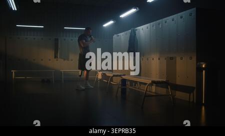 Junger Mann mit Prothesenbein wärmt sich auf und dehnt sich im sonnendurchfluteten Gym locker Room, nimmt eine Wasserflasche und bereitet sich vor, für intensive Trainingseinheiten zu gehen. Bestimmung und aktives Leben. Stockfoto