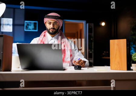 Ein Mann aus dem Nahen Osten in arabischer Kleidung sitzt am Tisch, vertieft mit Forschungen über seinen digitalen Laptop. Stockfoto