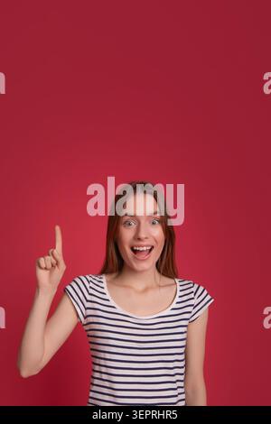 Aufgeregte Frau, die mit Zeigefinger auf rotem Hintergrund zeigt, mit Kopierraum, Idee, guten Nachrichten, Bildung, Beförderung, Lebensstil-Konzept für Schüler Stockfoto