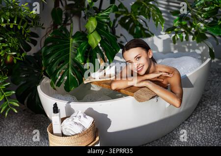 Glückliche weiße junge Frau, die sich in einem Sprudelbad in einem modernen botanischen Badezimmer entspannt Stockfoto