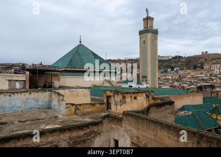 Die Al-Qarawiyyin-Moschee erhebt sich über verwitterten Medina-Dächern mit einem grünen Dach und einem kunstvollen Minarett unter einem bewölkten Himmel in Fes, Marokko. Stockfoto