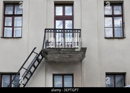 Fenster in dunklem Holz gerahmt, symmetrisch ausgerichtet. Rostiges Geländer deutet auf Alter und Verschleiß hin. Die Leiter lehnt sich an die Wand, was auf einen kürzlichen Zugang hindeutet Stockfoto