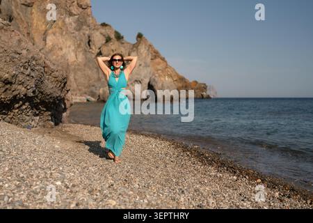 Frau, Strand, Sommer: Lächelnde Frau in türkisfarbenem Kleid spaziert am Kiesstrand an den Klippen des Ozeans unter klarem blauem Himmel. Stockfoto