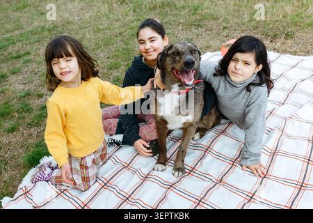 Drei Kinder sitzen zusammen mit ihrem Hund auf einer Picknickdecke in einem Park und schauen in die Kamera Stockfoto