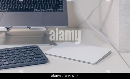 Ein Heimbüro mit einem Laptop auf einem Aluminiumständer, einer dunklen, kompakten Tastatur und einem weißen Trackpad auf einer sauberen, weißen Oberfläche, die eine gut organisierte Einrichtung für die Remote-Arbeit darstellen. Stockfoto