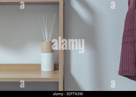 Ein weißer Reed-Diffusor auf einem Eichenregal, fotografiert neben einem bordeauxroten Strickkleid, das in der Nähe hängt und ein gemütliches und minimalistisches japanisches Zuhause mit warmer natürlicher Seitenbeleuchtung einfängt. Stockfoto