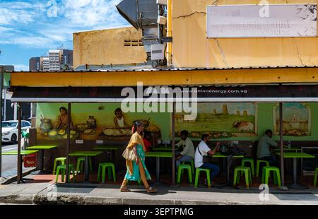 SINGAPUR - 29. JUNI 2025: Eine Frau läuft an einem lokalen Restaurant vorbei, in dem Gäste auf grünen Plastikhockern in einem Open-Air-Café in der Stadt sitzen Stockfoto
