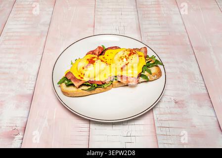 Offener Toast mit Speck, zwei pochierten Eiern, Hollandaise-Sauce und Paprika serviert auf einem weißen Teller über rosa Holz. Stockfoto