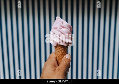 Minimalistischer Schuss einer Hand, die einen Schuss rosafarbenes Eis in einer Waffelkegel hält, positioniert auf einem auffälligen blau-weißen vertikal gestreiften Hintergrund, wodurch eine verspielte und grafische Sommerästhetik entsteht. Stockfoto
