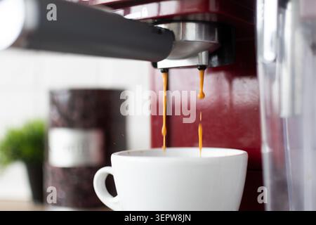 Nahaufnahme eines frisch gebrühten Espresso, der aus einer roten Kaffeemaschine in eine weiße Keramiktasse fließt. Stockfoto