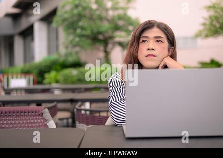 Thai Isan Transgender Geschäftsfrau in den 40ern, die nachdenklich mit Laptop im Freien wegschaut. Horizontales Mittelaufnahme-Porträt eines Reifen nicht-binären Southe Stockfoto