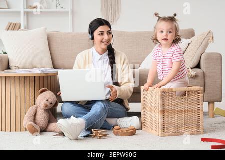Lächelnde Frau, die ferngesteuert mit Headset und Laptop arbeitet, während das Kleinkind zu Hause im Korb spielt, ferngesteuert arbeitet und die Mutterschaft ausbalanciert Stockfoto