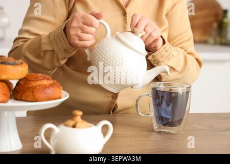 Seniorin gießt Schmetterlingserbsenblüten-Tee aus der Teekanne in eine Glasschale in der Küche Stockfoto