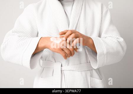 Junger Mann im Bademantel, der Creme auf der Hand vor weißem Hintergrund aufträgt, Nahaufnahme Stockfoto