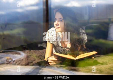 Junge Frau, die am sonnigen Fenster liest. Die junge Frau liegt auf einem Bett mit einem offenen Buch neben einem großen, sonnendurchfluteten Fenster, mit sanften Reflexen und Wärme Stockfoto