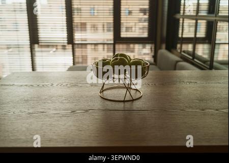 Frische Bio-Äpfel in einem Metallkorb auf einer Holzoberfläche angeordnet. Natürliches Licht filtert durch ein großes Fenster. Konzept des Wohnkomforts, Herbst Stockfoto