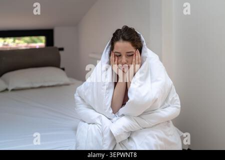 Traurige junge Frau, die auf dem Bett sitzt, in eine Decke gewickelt, das Gesicht auf die Hand gestützt und in Gedanken verloren hat. Begriff des Herzbruchs, Beziehungsschwierigkeiten, Lonelin Stockfoto