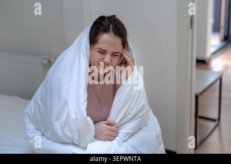 Unwohl Frau, in Decke gewickelt, sitzend auf dem Bett, berührend Schläfen, während sie an schweren Kopfschmerzen litt. Weibliche Grimassen vor Schmerzen und Unwohlsein, Conce Stockfoto