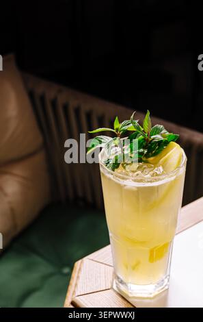 Ein mit einem gelben Getränk gefülltes Glas steht auf einem Holztisch in einem Café. Frische Zitronenscheiben und grüne Blätter ruhen darauf. Die Atmosphäre ist warm und einladend. Stockfoto