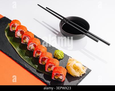 Eine Sushi-Platte sitzt auf einem schwarzen Schieferbrett. Die Sushi-Brötchen werden mit leuchtendem orangenem Fischrogen belegt. Eingelegter Ingwer und Wasabi sitzen neben den Brötchen. Stäbchen ruhen neben der Schüssel mit Sojasauce. Stockfoto